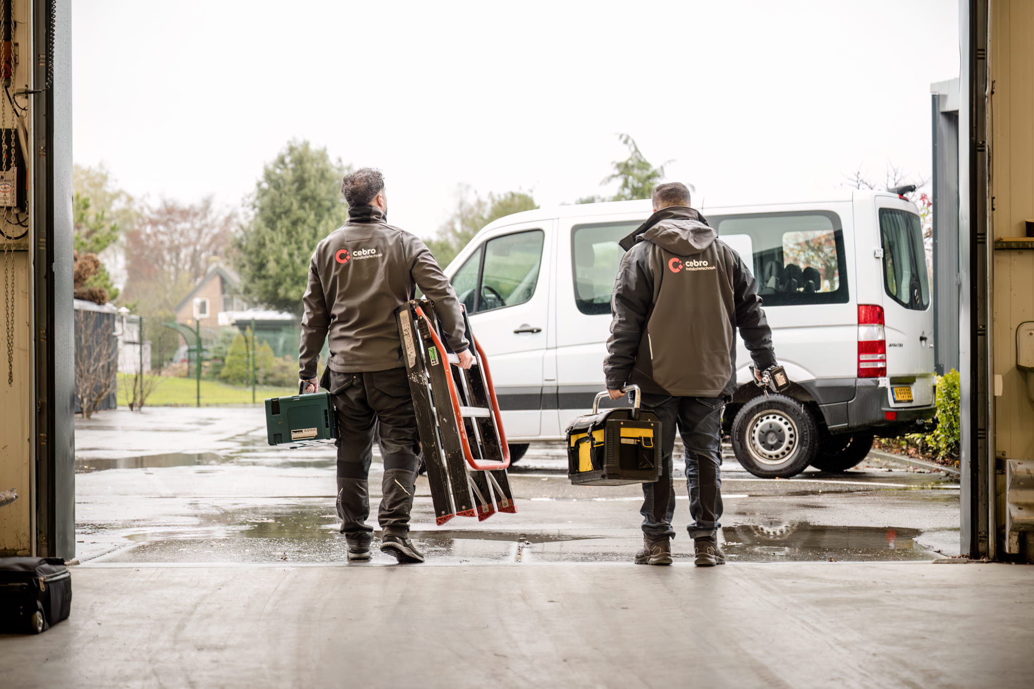 Two workers in matching jackets walk out of a loading bay carrying a ladder and a toolbox toward a white van in a residential, wet-yard setting.