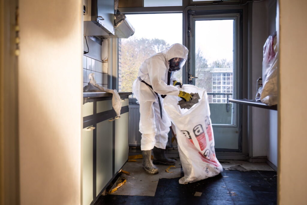 Hazmat-suited worker collects debris into a large white bag in a sunlit kitchen-like room.